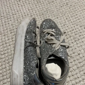 Glittriga silvriga sneakers från Tamaris - Snygga sneakers från Tamaris i storlek 37, täckta med silverglitter och med vita snören. Otrolig detalj till en basic outfit.