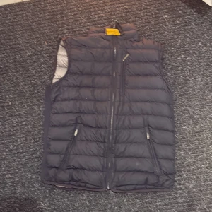 Svart dunväst från Parajumpers  - Säljer en svart dunväst från Parajumpers i storlek YL. Västen är quiltad och superlätt med dragkedja framtill och två snedställda fickor med dragkedja. Hög krage och klassisk Parajumpers-detalj i nacken. Perfekt för lager-på-lager under kyliga dagar. Skick 8/10