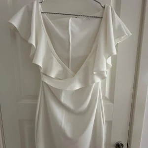 Vit volangklänning från NLY One - Använt endast en gång till student. Superfin vit kortklänning från NLY One med v-ringning och stora volangdetaljer över axlarna. Klänningen har en figurnära passform och är tillverkad i ett mjukt, slätt material. Perfekt för dig som vill sticka ut med en elegant och stilren look. 