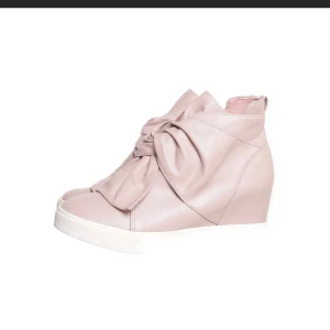 Rosa sneakers med kilklack och rosett - Trendiga sneakers i ljusrosa skinnimitation med vit sula och kilklack. Skorna har en stor dekorativ rosett framtill som ger en unik och cool look. Dragkedja på sidan för enkel påtagning. Perfekta för dig som vill sticka ut med din stil.