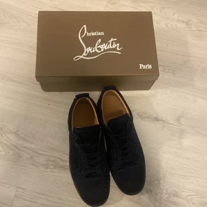 Loubs - Snygga svarta sneakers från Christian Louboutin i mocka med klassisk röd sula. Skorna har rund tå, svarta snören och diskret design som passar perfekt till streetwear. Insidan är i läder för extra komfort. Kommer med originalkartong och tillbehör.