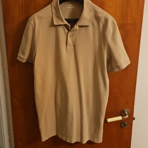 Beige pikétröja från Dressmann L - Säljer en stilren beige pikétröja från Dressmann i storlek L. Klassisk krage, knäppning med två knappar och korta ärmar. Skön bomullskänsla och passar perfekt till jeans eller shorts för en clean look.