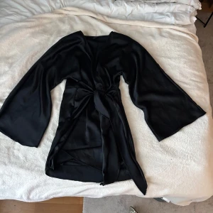 Svart omlottklänning med vida ärmar - Snygg svart kort klänning i satinliknande material med breda, vida ärmar och knytning i midjan. Klänningen har en v-ringning.