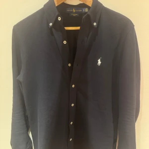 Mörkblå skjorta från Ralph Lauren - Snygg mörkblå skjorta från Ralph Lauren i featherweight mesh-bomull. Klassisk button-down krage, vita knappar och ikonisk broderad logga på bröstet. Långärmad och normal passform, perfekt till jeans eller chinos.