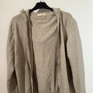 Beige hoodie från The Tailoring Club - Beige hoodie från The Tailoring Club i mjukt material. Tröjan har dragkedja framtill, huva och långa ärmar. Perfekt för en chill och avslappnad stil. Passar dig som gillar neutrala färger och enkel design.