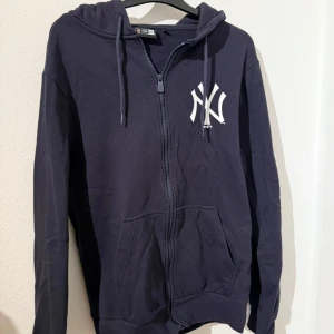 Mörkblå Yankees hoodie med dragkedja - Mörkblå hoodie från New Era med New York Yankees-logga på bröstet. Tröjan har huva med snörning, dragkedja framtill och två stora fickor. Perfekt för dig som gillar sportig och avslappnad stil.