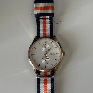 Regal klocka med randigt band - Stilren klocka från Regal med vit urtavla och roséguldfärgat boett. Bandet är randigt i marinblått, vitt och orange, vilket ger en sportig och fräsch look. Urtavlan har minimalistiska index och två mindre urtavlor för extra funktion.