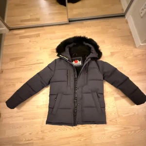 Grå/svart Zavetti Canada - Snygg svart vinterjacka från Zavetti Canada med luva och svart päls. Jackan har flera fickor, dragkedja framtill och coolt märke på ärmen. Perfekt för kalla dagar när du vill hålla stilen och värmen.