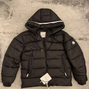 Svart dunjacka från Moncler med huva - Svart dunjacka från Moncler med huva och vita detaljer. Jackan har quiltad design, dragkedjor på fickorna och framtill samt en klassisk Moncler-logga på ärmen. Perfekt för kalla dagar när du vill hålla stilen och värmen. Passar även S
