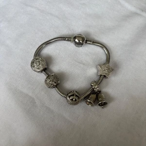 Pandora armband med berlocker i silver - Snyggt Pandora-armband i silverfärgad metall med flera glittriga berlocker, bland annat en stjärna, klockor och peace-symbol. Armbandet har detaljer med stenar och olika former som ger en cool och personlig look.