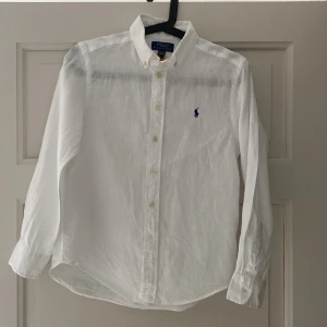 Vit linneskjorta från Polo Ralph Lauren barn/kille  - Klassisk vit linneskjorta från Polo Ralph Lauren. Skjortan har lång ärm, knappar framtill och ikoniska broderade loggan på bröstet. För barn/kille storlek M (10-12)