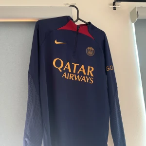 PSG träningsbyxor från Nike, marinblå - Snygga marinblå PSG träningsbyxor från Nike med guldfärgade detaljer och klubbmärke på låret. Byxorna har smal passform, elastisk midja och coolt mönster på benen. Perfekta för fotbollsträning eller chill. 