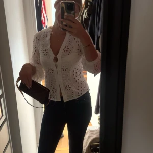 Vit broderad blus med volang - Superfin vit blus med broderade hålmönster och volangkant längs v-ringningen. Blusen har trekvartsärmar och knappar framtill, samt en lätt croppad passform. 