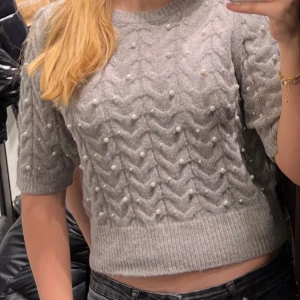 Grå stickad tröja med pärlor från Zara - Superfin grå stickad tröja från Zara med kort ärm. Tröjan har ett snyggt zigzag-mönster och är dekorerad med små pärlor över hela framsidan. Perfekt att styla med jeans eller kjol för en trendig look. Endast använd 1 gång, den ser ut som ny, storlek M men passar bra på mig som brukar ha Xs och S💕