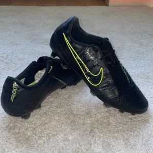 Nike Phantom Venom fotbollsskor svart/neon - Säljer ett par NIKE PHANTOM VENOM ELITE FG UNDER THE RADAR - SVART/NEON fotbollsskor i svart med neongula detaljer och holografisk Swoosh. Skorna har snörning, texturerad ovandel för bättre bollkontroll och dobbar för spel på gräs. Snygg, modern design som sticker ut på planen. Skriv vid frågor eller andra funderingar.        Mvh Axel 
