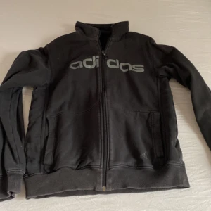 Svart ziptröja från Adidas - Svart Adidas ziptröja med stor logga över bröstet och snygg dragkedja med rund detalj. Tröjan har höga muddar, två fickor fram och är tillverkad i mjuk bomullsmix. Perfekt för chill dagar eller till träningen.