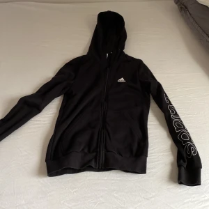 Svart Adidas hoodie med dragkedja - Svart hoodie från Adidas med vit logga på bröstet och stort Adidas-tryck längs ena ärmen. Tröjan har huva, två fickor fram och hel dragkedja. Materialet är mjuk bomull och polyester, perfekt för chill eller träning. Klassisk och clean stil som funkar till allt.