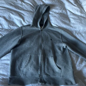 Grå hoodie från Polo Ralph Lauren - Säljer en grå hoodie från Polo Ralph Lauren med dragkedja och huva. Tröjan har långa ärmar, två fickor fram och en liten broderad logga på bröstet. Perfekt för chill dagar och enkel att matcha med jeans eller joggers.
