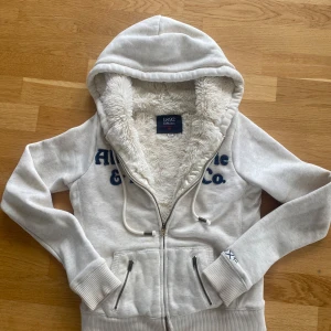 Vit hoodie Abercrombie & Fitch pälsfodrad - Supermjuk vit hoodie från Abercrombie & Fitch 1892 Collection med fluffigt pälsfoder, broderad logga i mörkblått, dragkedja, snörning i huvan och två fickor med dragkedja. Perfekt för kyliga dagar och riktigt cozy vibes.