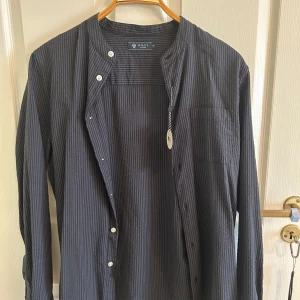 Randig blå henley från Riley - Snygg randig henley-skjorta från Riley i mörkblått, tillverkad i luftig bomull. Skjortan har ståkrage, knappar framtill och en bröstficka. Perfekt för dig som gillar stilren och avslappnad look. Köptes för 699kr