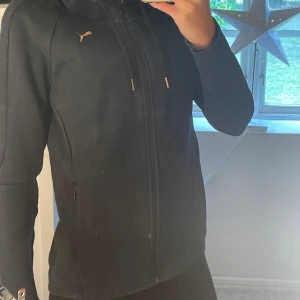 Svart sportig jacka från Puma - Svart jacka från Puma med dragkedja hela vägen, huva och snygga gulddetaljer på snörningen. Jackan har en slim och sportig passform, perfekt för dig som gillar en clean och stilren look. Puma-logga i guld på bröstet och praktiska fickor med dragkedja.