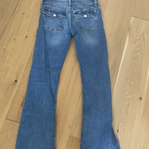 Blå bootcut jeans med slitningar - Säljer ett par blå bootcut jeans med snygga slitningar på knäna och klassiska bakfickor med lock och knapp. Jeansen har normal passform och är tillverkade i denim med en cool, avslappnad vibe.