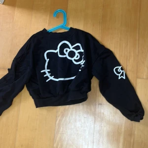 Svart Hello Kitty croppad tröja - Svart croppad tröja med stor vit Hello Kitty-tryck på ryggen och tryckta detaljer på ärmarna. Tröjan har långa ärmar och ribbad nederkant. Perfekt för dig som gillar streetstyle och vill sticka ut med en söt men edgy vibe.