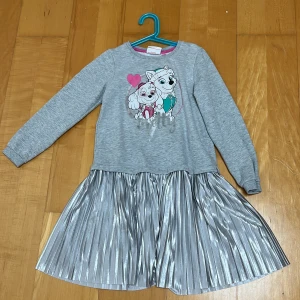 Grå klänning med Paw Patrol och silverkjol - Supersöt grå klänning med långärmad överdel och tryck av Paw Patrol-karaktärer framtill. Kjolen är plisserad och skimrande i silver, vilket ger en cool kontrast. Perfekt för dig som gillar både bekvämt och glittrigt! Rund hals och mjukt material upptill.