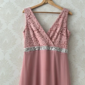 Lång rosa spetsklänning med paljetter - Säljer en elegant långklänning i ljusrosa med v-ringad omlott-topp i spets och blommigt mönster. Midjan är dekorerad med ett brett band av silvriga paljetter. Kjolen är mjukt fallande och klänningen är ärmlös, perfekt för bal eller fest.använd endast 2 timmar köpt från Lindex nypris 600kr 