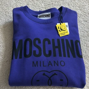 Blå/lila Moschino sweatshirt med tryck - Snygg blå/lila sweatshirt från Moschino med stort svart logotryck och Milano-text på bröstet. Tröjan har rund hals, långa ärmar och en relaxed passform. Perfekt för dig som gillar statement-plagg och vill sticka ut med ett ikoniskt märke.