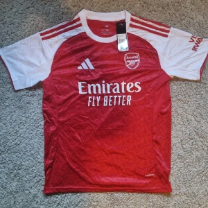 Arsenal 26 Tröja - Arsenal hemmatröja från Adidas i rött och vitt med klubbmärke och sponsortryck på bröstet. Klassisk design med vita ärmar, röda axelränder och subtilt mönster i tyget. Tillverkad i lätt och ventilerande Aeroready-material, perfekt för match eller träning.
