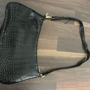 Svart axelväska i croco-imitation - Snygg svart axelväska med croco-mönstrad yta och guldfärgade detaljer. Knappt användt. Har ett litet snöre som sticker ut men går att klippa!