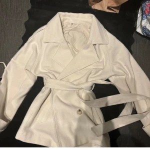 Vit trenchcoat med bälte - Snygg vit trenchcoat med bred krage och knytskärp i midjan. Jackan har dubbelknäppning och är tillverkad i ett mjukt tyg som ger en clean och stilren look. Perfekt för dig som vill ha en klassisk och trendig jacka till garderoben.