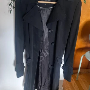 Svart trenchcoat med bälte - Stilren svart trenchcoat från Rocky LM med klassisk krage, stora knappar och bälte i midjan. Jackan har långa ärmar med snygga detaljer vid ärmsluten och fickor framtill. Perfekt för dig som vill ha en tidlös och clean look.