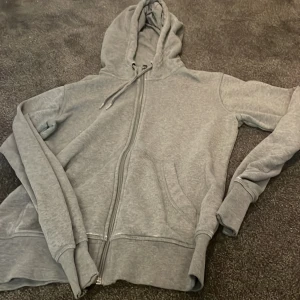 Grå hoodie från Essentials med dragkedja - Säljer en grå hoodie från Essentials i storlek S. Tröjan har huva med snörning, dragkedja framtill och två fickor. Perfekt för chill dagar eller när du vill ha en enkel och clean look. Materialet känns mjukt och skönt mot huden.