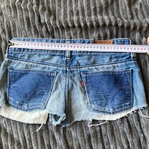 Levi's jeansshorts med rå kant - Snygga blå jeansshorts från Levi's med klassiska bakfickor och rå, fransig kant. Shortsen har låg midja och en tight passform som sitter snyggt på kroppen. Perfekta för varma sommardagar och har den ikoniska röda Levi's-taggen på bakfickan.