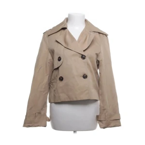 Beige kort trenchcoat från H&M XS - Kort trenchcoat från H&M i beige med dubbelknäppning och bred krage. Jackan har snygga detaljer vid ärmslut och axlar, samt stora knappar framtill. Tillverkad i bomull och polyamid med polyesterfoder. Perfekt för dig som gillar klassisk stil med modern twist.