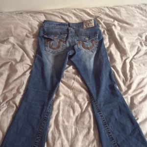 True Religion Joey Low Rise Flare Jeans - Säljer ett par blå True Religion Joey jeans med låg midja och flare-ben. Klassiska kontrastsömmar och stora broderade bakfickor med logga. Jeansen är i denim med snygg tvätt och detaljerade nitar. Kan tänka mig byta mot et par liknade. Pris kan diskuteras, köpte för 2300