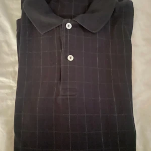 Mörkblå rutig pikétröja Polo Ralph Lauren - Snygg mörkblå pikétröja från Polo Ralph Lauren med diskret rutigt mönster och klassisk krage. Tröjan har knäppning med två knappar framtill och är perfekt för en stilren look. Materialet känns mjukt och bekvämt mot huden.