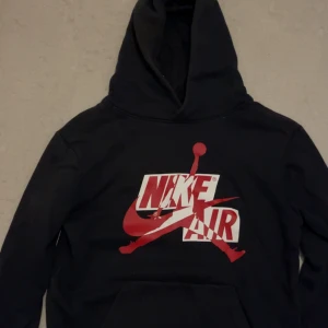 Svart Nike Air Jordan hoodie med röd logga - Svart hoodie från Nike Air Jordan med stor röd och vit logga på bröstet och klassisk Jumpman-figur. Tröjan har huva och magficka, tillverkad i mjuk bomullsmix. Perfekt för dig som gillar streetwear och sportig stil.