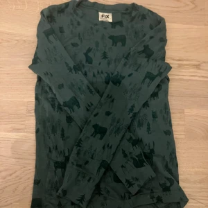 Grön långärmad merinotröja från FIX - Säljer en mörkgrön långärmad tröja från FIX i ren merinoull. Tröjan har ett coolt djur- och skogsmönster med björnar, rådjur och träd över hela plagget. Perfekt för dig som gillar natur och vill ha något mjukt och skönt på dig. För barn!🙃