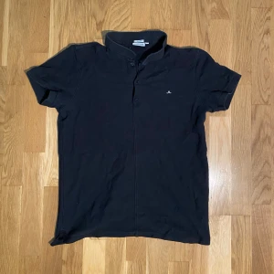 J.Lindeberg piké - Svart t-shirt från J.Lindeberg i slim fit-modell med korta ärmar och klassisk krage. Diskret logga på bröstet och knappslå framtill. Materialet är mjuk bomull som känns skönt mot huden. Perfekt för dig som gillar stilrena och enkla plagg. Storlek M men passar bättre S.