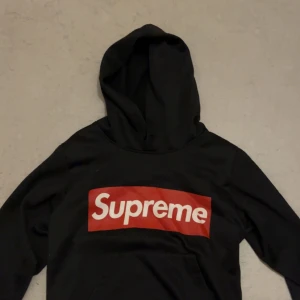 Supreme svart hoodie med röd logga - Supreme hoodie i svart med den klassiska röda boxloggan framtill. Tröjan har huva och långa ärmar, tillverkad i mjuk bomull. Perfekt för dig som gillar streetwear och vill sticka ut med en ikonisk look.