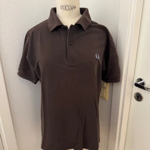 Brun Fred Perry piké  - Snygg brun piké från Fred Perry i 100% bomull. Klassisk design med krage, korta ärmar och två knappar framtill. Fred Perry-logga. Stl S. Sitter på en dam-docka på bilden. Hämtas gärna på Hägerstensåsen i Stockholm för fraktfritt. 