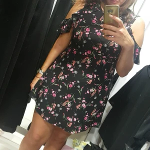 Svart blommig kortklänning med volang - Supersöt svart kortklänning med blommigt mönster och korta ärmar med cut-out volangdetaljer. Klänningen har en lätt A-linjeform och är perfekt för dig som gillar romantiska och trendiga plagg. 