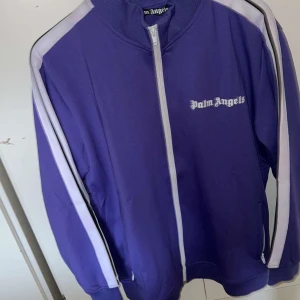 Palm Angels Tracksuit Lila  - Säljer en snygg tracksuit från Palm Angels i lila med vita sidoremsor och broderad logga på benet. Den passar för storlek L kanske M. Den är i Nyskick då jag inte har använt den och Fri frakt ingår