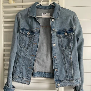 Ljusblå jeansjacka från Bershka - Snygg ljusblå jeansjacka från Bershka med klassisk krage, silverfärgade knappar och två bröstfickor med lock. Passar både xs och s