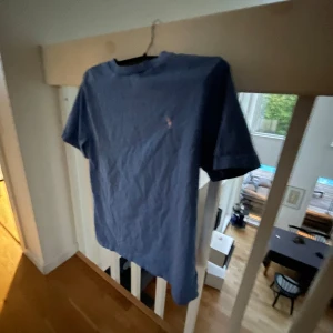 Blå t-shirt från Ralph Lauren - Snygg blå t-shirt från Ralph Lauren med klassisk rund hals och korta ärmar. På bröstet finns det ikoniska broderade Polo-loggan i rosa. T-shirten är gjord i mjuk bomull och har en clean, enkel look som funkar till jeans eller shorts.