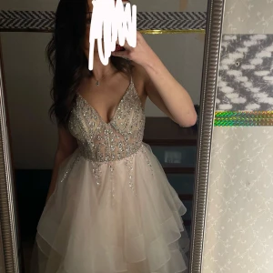 Balklänning med paljetter - Elegant champagnefärgad balklänning med glittrande paljetter och volanger. Klänningen har en v-ringad design och är ärmlös med en lång, flödande kjol. Perfekt för en glamorös kväll!