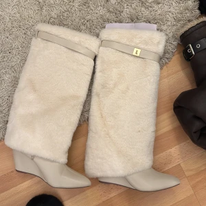 Beige stövlar med pälsdetaljer - Snygga beige stövlar med mjuk pälsdetalj och kilklack. Perfekta för att hålla värmen under kyliga dagar. Stövlarna har en dekorativ rem med gulddetalj upptill.